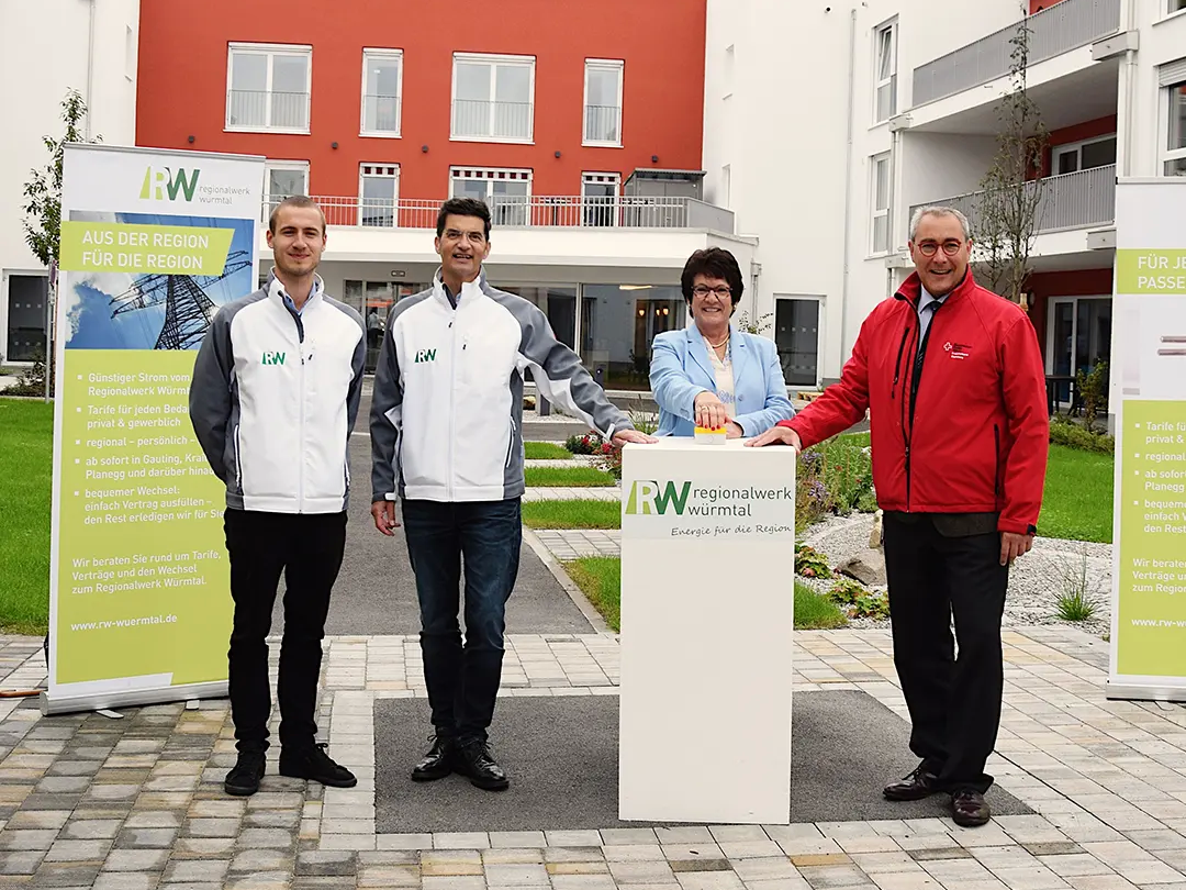 Vier Personen stehen vor dem Mehrgenerationen-Campus in Gauting. Sie posieren gemeinsam um ein weißes Podest mit dem Logo des "RW Regionalwerk Würmtal". Im Hintergrund sind Werbebanner zu sehen.